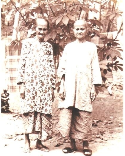 This is a photo of my great-grandmothers, sisters Aminah (left) and Siah (right) Binte Abdul Rahmanâor more affectionately known as Tok Aminah and Tok Siah. Tok Aminah was my grandmother's mother, whereas Tok Siah was my grandfather's.
Tok Aminah resided in Kampung Sedili Besar, Johor, whereas Tok Siah was in Kampung Pahang in Pulau Tekong Besar, Singapore. This photograph was taken during one of their visits to Pulau Ubin in my grandparents' courtyard.
According to my mom, both Tok Aminah and Tok Siah enjoyed consuming daun sirih (betelnut leaves) together with kapur sirih (limestone paste), gambir (Uncaria gambir), and buah pinang (Areca catechu). Sometimes, the leaves and ingredients would be eaten as-is together, or they would be refined together using an apparatus known as 'gobek sirih'.
Tok Siah in particular was renowned for her 'sentil tembakau' âa traditional Malay method of consuming tobacco, where a pinch of shredded tobacco would be 'brushed' against the teeth and later tucked between the gums and cheek. Any liquid extracts, presumably a mixture of tobacco and saliva, would be spit out. My mom recalled how Tok Siah would tuck her sentil tembakau into random spots, like window panes, whenever it was time for her daily obligatory prayers, only to retrieve it and continue using it afterward. She was never without it.
Additionally, both sisters frequently wore terompahâa traditional wooden clogâwhile in their rumah dapur (kitchen annexes) or outside when they were collecting water from the well. My mom remembered hearing their steps making a distinct "ketak ketuk" sound on the ground.
Tok Aminah passed away in the mid-1970s in Kampung Sedili Besar, while Tok Siah passed away on 26 November 1985 in her fifth child's 3-room HDB flat in Serangoon, just briefly after their eviction from Pulau Tekong Besar to mainland Singapore.