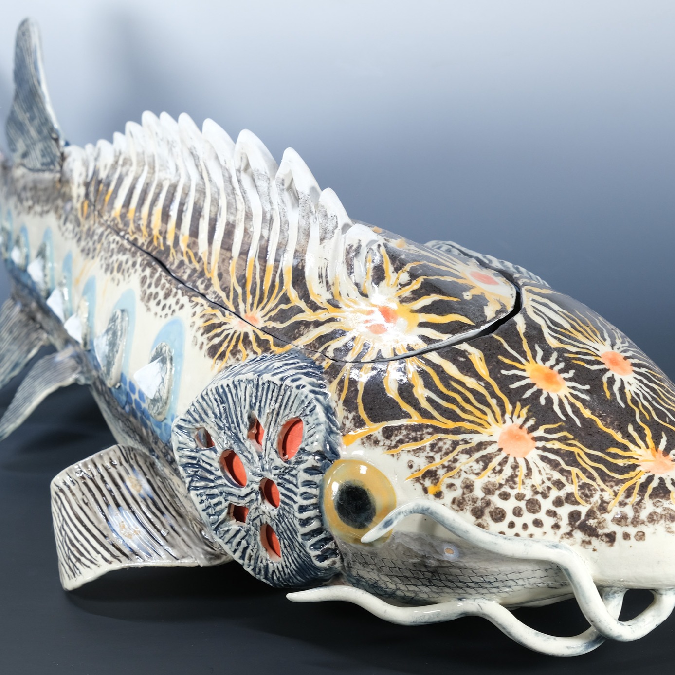 Cosmic Sturgeon, 2025
Ceramic lidded vessel
7.5"H x 10"W x 26"D
This sculpture is a personal narrative about finding comfort in knowing that we are all connected by nature and the cosmos.
Fresh from the kiln, it has been accepted into the @the.groundhog.show in Akron, OH. This annual exhibit supports queer culture and gives space and solidarity to the LGBTQIA+ arts community in Northeast Ohio. Proud to be included!
Here is the short version of the long story behind this artwork:
I was visited by a sturgeon on Lake Michigan while caring for my mother during her last battle with ovarian cancer. After strawberry picking in Algoma, WI, the lake was calling me to visit. I found a bench at a dead-end that overlooked a rocky incline to the shores. Basking in the sun, eating fresh-picked strawberries, enjoying a moment of alone before returning home.
Staring into the sparkling water, a gigantic lake sturgeon appeared. We stared into each otherโs eyes for what seemed like hours. They reminded me that we are all nature, that we all return to the cosmos at some point, and comforted me. After my fill of comfort and strawberries, I decide to ascend the rocky cliff to the small sandy shore.
A few steps down, a badger popped out of his hole, both of us surprised, side-eyed each other, and poised to run. Slowly backing away, I decided it was best to leave.
A few days later, my mother and I took a car ride to get out of the house. Enjoying the winding country roads with moments of silence that eased hard questions, taking the conversation to further depth. We ended up on the same lakeside road near Algoma, so I brought her to the magical bench and we looked for the sturgeon while holding hands.
Neither the sturgeon, or badger, made an appearance and the sun was too strong for her cancer drug sensitive skin to wait very long. We went home and made strawberry jam.
My mother was my first home, unconditional love, and being who gave me life. Holding hands through it all, together, even when we lived apart on earth. Through nature, through the cosmos, we never have to let go.
#ceramicsculpture #sturgeon #vessel