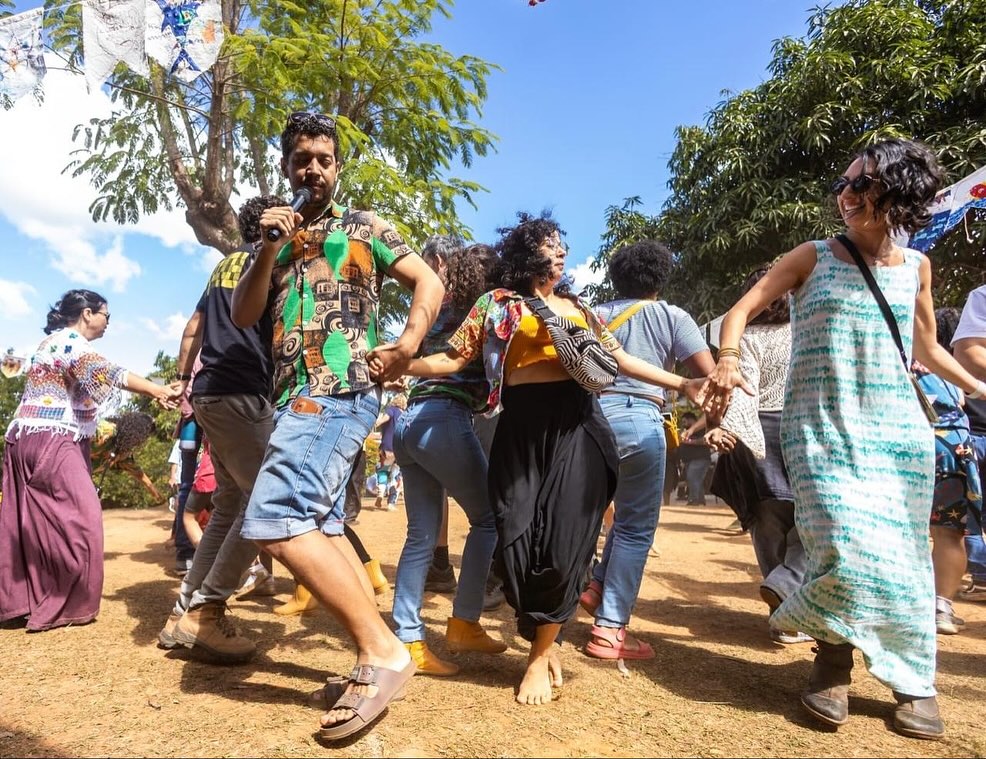 Nunca vamos nos esquecer da sua primeira visita por aqui: “olha, eu sou um homem preto, gay e pai solo. Meu filho já viveu muitas violências e eu preciso de um lugar seguro para ele e também para mim”. Foi assim que Bruno se apresentou. Um sujeito marcado pela trajetória de ser quem se é, com consciência da sua existência e orgulhoso de sua identidade, chegou. Chegou no lugar certo! Iniciou um projeto denominado “Democracia em Movimento” com os adolescentes da escola, com o objetivo de reforçar a importância da participação juvenil em ações de cidadania. Colaborou na aproximação de diálogos com lideranças indígenas Yanomami e trouxe muitas reflexões potentes sobre justiça social, visto que era um Assistente Social engajado e comprometido atuando no Ministério do Desenvolvimento Social.
No domingo à noite, recebemos a difícil notícia que ele havia sofrido um afogamento no Rio Negro, e ao longo da semana foi confirmada sua partida. Por aqui, lidando tanto com a vida, temos que aprender a lidar com a morte, com a realidade que não é escrita com enredos planejados. Essas fotos são os registros da nossa Festa do Campo desse ano. Bruno foi designado para puxar todo o grupo na dança, e assim fez com alegria e orgulho. Vamos seguir aqui no compromisso sincero de cuidar, educar, acolher, chorar junto de seu filho e reafirmar que escola é lugar do comum, de comunidade, de sentir junto, sentir tudo.
Ficamos com o carinho e parceria da última mensagem enviada por ele para a direção pedagógica, no dia da abertura do seminário de 10 anos da escola.
“Ei Letícia, bom dia! Passando pra desejar um bom evento, que os encantados deem caminho para que tudo corra bem. Minha agenda está bem apertada, mas darei um jeito de passar pra prestigiar a escola e o legado que ela deixa nas relações dos nossos filhos. Exú dê caminho!”