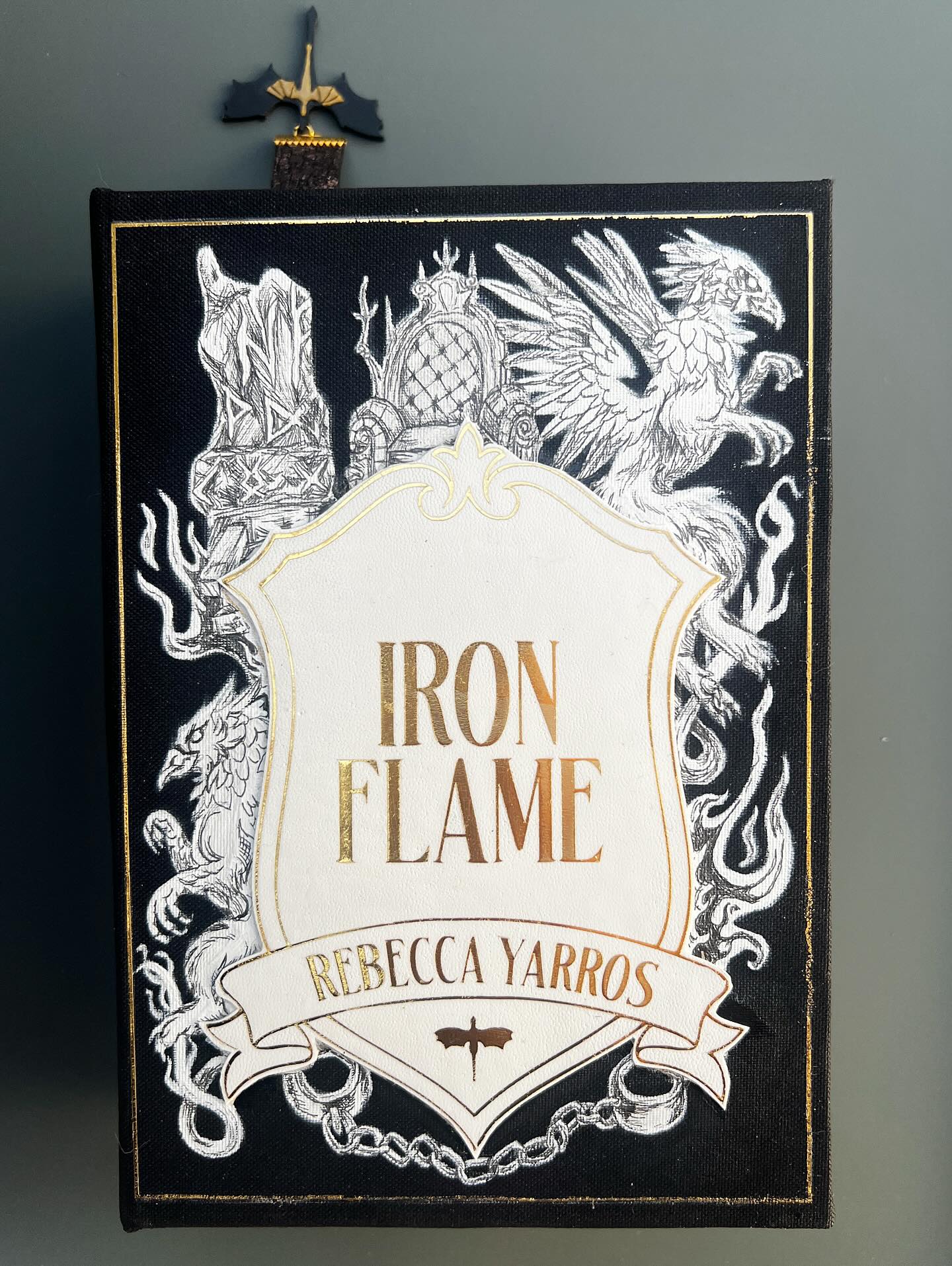 Full breakdown for the Iron Flame rebind, 🚨🚨🚨 spoilers abound!
01. The cover of each of these books features a simple shield with the title and author name surrounded by illustrations of prominent characters and scenes from the book. Fans will notice the throne, griffins, and chains used to restrain Violet during her grueling interrogation to name a few.
02. Violet begins harnessing her signet in this book and a special saddle is crafted to help her keep her seating on Tairn which works uh… sometimes. I used gouache to capture the image of her losing her seat and dropping her knives through the sky as her black morningstar scoops to catch her.
03. Each book’s spine takes on the look of a Basgiath textbook on the shelf.
04. Speaking of a black morningstar tail, take a look at this fine fellow cutting through the air. Each back cover features a portrait of a prominent dragon with a shield emblazoned with the war college’s name as an emblem.
05. This book’s inscription is a beautiful line from Andarna which represents why she picked her after decades in her egg: “You would have the heart of a rider and the mind of a scribe. You would be mine.” For this series, I custom printed the endpapers with a thundercloud motif that I used a gold pen to hand craft lightning bolts and letter the verbiage to mimic strikes across the page.
05. Another shot of Tairn catching Violet Sorrengail out of the sky.
06. The hero art on the back cover, war college’s textbook spine, and bookmark with 3D printed earring bookmark charm.
07. More details 🖤🐉⚡️
