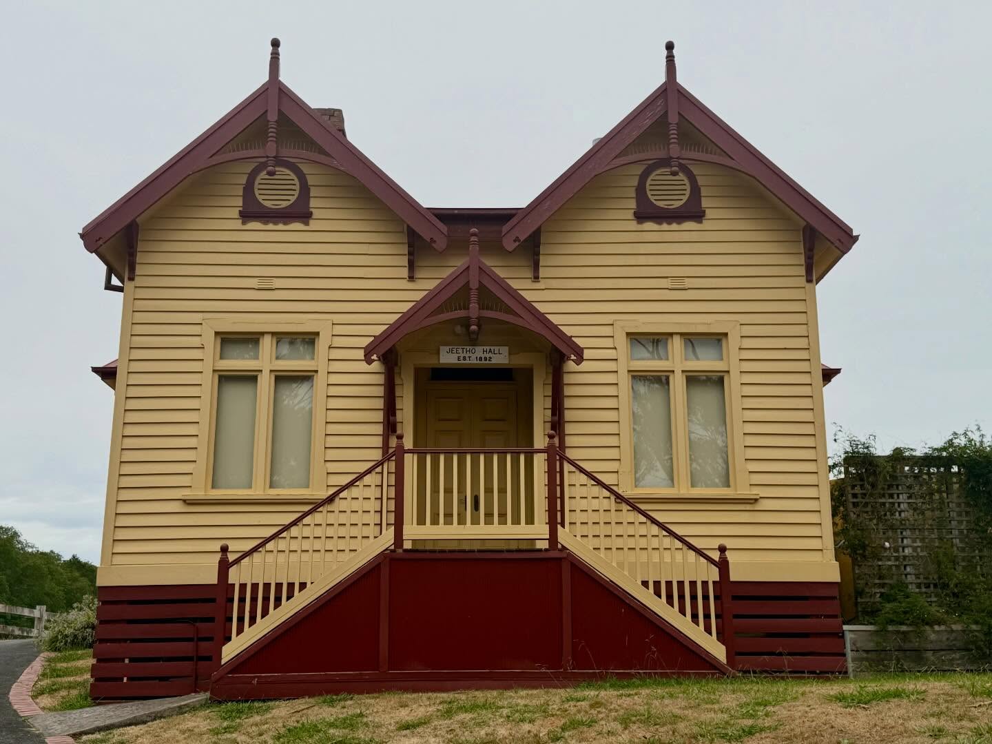 Former Shire of Poowong and Jeetho Council Chambers, Jeetho Valley, South Gippsland (1892) (HO46, South Gippsland Shire). A fieldwork find with a storybook silhouette. The township it was built to serve largely dissipated as nearby centres expanded on the back of coal mining, yet a small rural community has cared for the place since 1908.
The now little-known Melbourne architect John Waugh is credited with its picturesque, paired steep-gabled composition and Carpenter Gothic inflection, including king-post-like drops to the finials/pendants and oculus (round) louvred vents. These are motifs more typical of ecclesiastical buildings than council chambers, and correspondingly uncommon at a civic scale.
Waugh seems to have been peripatetic: associated with the PWD in Tasmania, later active in Subiaco (WA), and in Sydney by the late Federation era. The final two images capture the building’s quiet, back-country setting.