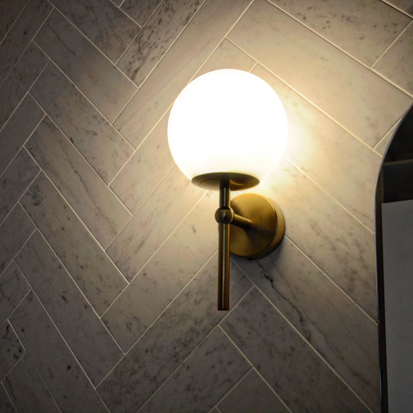 Natural Carrara marble in a herringbone pattern, lit by a warm brushed gold wall light for timeless bathroom style installed in one of our duplex builds.