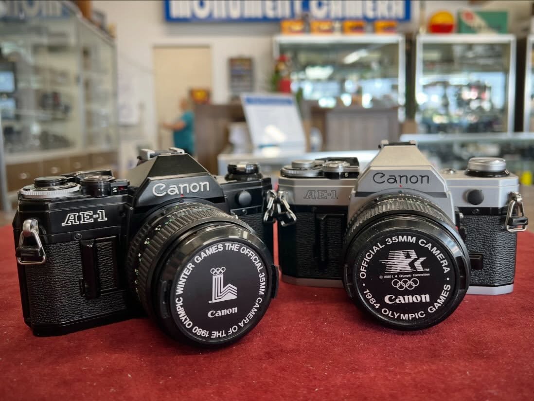 Early 1980s Canon AE-1s, black and silver bodies, with commemorative Olympics lens caps corresponding to their manufacture dates. #canonae1 #canonfilmphotography #canonslr #camerastore