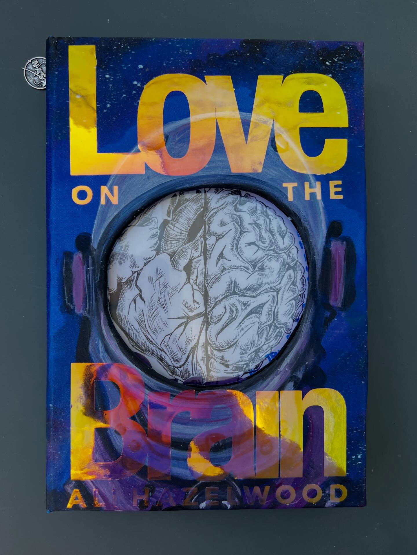 The full breakdown of my rebind of Love on the Brain by @alihazelwood Light spoilers and hardcore fangirling + special interests ahead.
01. At age 7, I was convinced I would become an astronomer and now I’m a graphic designer so to say I’ve seen the NASA brand guidelines a time or two would be putting it lightly. For the cover I went with a blend of Helvetica (the NASA typeface) and Avenir (something I’ll weave into - spoiler - future rebinds of other books by her to build some consistency). The story involves two perceived adversaries working on a revolutionary helmet (BLINK) for astronauts that involves brain stimulus in space so I made a cut out and made a faux viewport of the helmet into the rest of the novel.
02. The typography was done in duochrome, translucent HTV so that from certain angles/lighting it totally sticks out and in others it completely vanishes. It’s one of my favorite materials to work with and added a cool effect to what was otherwise a very minimal, connected color palette throughout the whole piece.
03. I hunted down a NASA logo charm for the bookmark on Etsy. It’s obviously the meatball version and not the spaghetti one because, again, my special interests really collided on this one.
04. The inside endpapers have a split design, printed text, and use a sheet of transparency (all firsts for me). The front cover is a hand-painted galaxy motif that echoes the color of Bee’s hair and space vibes. For a STEMinist novel, I came up with the folded transparency trick to both be a sort of windshield from the front and also have handwritten quotes like an old-school transparency for a projector. Very✨academia✨
04. A shot showing all of the layers and the anatomical drawing of Love (heart) 🫀on the Brain. 🧠 and accurate labels for the parts of each.
05. The back endpapers feature my take on the sketch Bee drew for Levi to explain how she would address the motherboard issue which she writes parts of in German.
06 and beyond: more detail shots of the pink/purple Bee-hair gradient, artistic license taken with the calico fur of Felicette, and meeting the author herself so show her my insanely detailed love letter to the book.