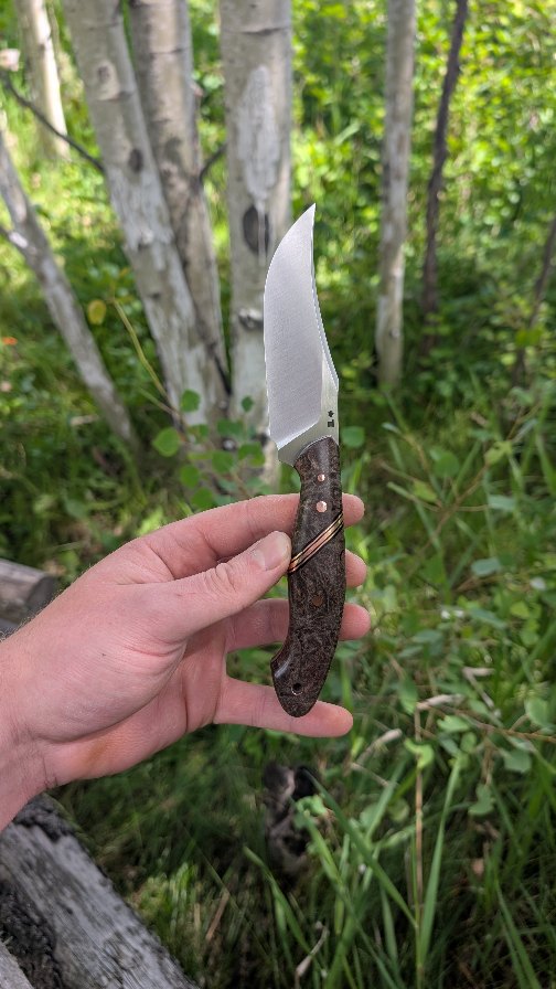 Every part of the knives I make are custom, from the handle material and steel finish, to even the style of the blade. When the client reached out, he was looking for an exaggerated clip point with the handle of my Mesa line. That isn't something I offered at the moment, so after talking about specifics and sharing a few drawings, we settled on a design that worked for the two of us. Making everything myself from scratch, means I can easily alter designs on the fly.
The handle and blade finish are also his choice working within the materials I had. The request of "black wood with some grain variety" immediately made me think of my block of black dyed maple burl. Sharing more photos and drawings, the black maple burl tied with the copper hardware and segment made for a beautiful, functional, and stylish handle.
Copper is beautiful, but so tricky to work with. Not only does it get extremely hot very quickly from the friction of grinding, but it naturally grinds slower than the surrounding wood making a "copper speed bump". Therefore, precautions and attention to detail must be kept to keep the handle strong and the scales smooth, perfectly flush, and polished.
Just because it's made for use doesn't mean it can't be beautiful.
Statistics.
Style: Clip Point Mesa
Steel: NitroV
HRc: ≈62
Overall length: 8.75"
Blade length: 4"
Cutting edge: 3.75"
Stock: ⅛"
Handle: Black Maple Burl, Copper, G10
Liner: Black G10
Pins: Copper
Tube: Copper
As always, every step from profiling, to heat treat, to polishing, to leather work is all done myself in Northern BC, Canada.
Go off the edge of your trail.