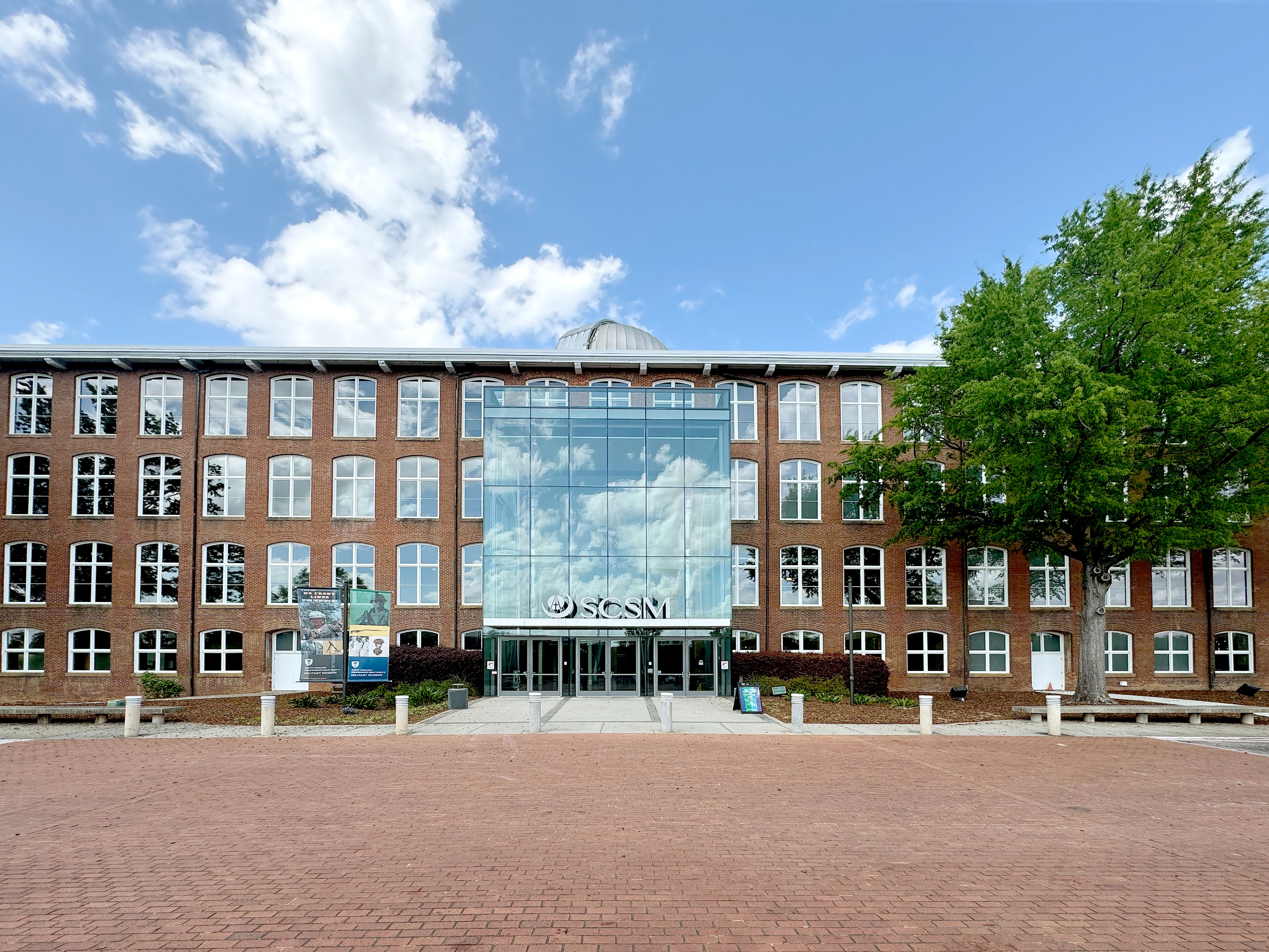 2026 South Carolina Historic Preservation Award Spotlight: South Carolina State Museum for the Columbia Mills Building
We are proud to celebrate the South Carolina State Museum as a recipient of a 2026 South Carolina Historic Preservation Award for Heritage Tourism, recognizing the thoughtful rehabilitation of the Columbia Mills Building!
Built in 1893 and expanded in 1901, the Columbia Mills Building is one of South Carolina’s most significant industrial landmarks. Designed by Lockwood, Greene, and Company, the four-story brick mill played an important role in the state’s textile industry and is widely recognized as one of the first textile mills in the United States powered by electricity.
Listed in the National Register of Historic Places in1982, the building now serves as the home of the South Carolina State Museum, bringing this remarkable piece of industrial history to thousands of visitors each year.
The recent rehabilitation addressed critical building-envelope issues while carefully respecting the character of this historic resource. Work included new roof systems, complete replacement of non-historic windows, improvements to the atrium enclosure, and repairs to deteriorated eaves and rafter tails.
The new custom aluminum windows are a particularly thoughtful example of preservation in practice. Guided by historic imagery, the designs replicate the scale, proportions, and profiles of the building’s original industrial windows while providing the durability and performance needed for contemporary use.
At the atrium, a new roof design with dormers preserves the original intent of bringing natural light into the space while significantly improving weather protection and reliability. The rehabilitation also expanded and reconnected the greenhouse to the atrium, creating a more functional gathering and event space for the public.
This project demonstrates that preservation and modern performance can work hand in hand. Through careful research, collaboration, and sensitive design, the project team extended the life of a nationally significant landmark while ensuring that it can continue serving South Carolinians and visitors.