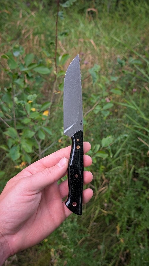 It's been quite some time since I've made a Kindling 4XL and it was great to get back to one of my original designs. With a 4" blade and a 4.75" handle, it fits comfortably in XL hands with plenty of blade length to get work done. The black burlap is sleek, but has some incredible depth to it while there is a great colour pairing of the canvas liners and copper hardware.
Just because it's made for use doesn't mean it can't be beautiful.
Statistics.
Style: Kindling 4XL
Steel: NitroV
HRc: ≈62
Overall length: 8.75
Blade length: 4 ⅛"
Cutting edge: 4"
Stock: ⅛"
Handle: Black Burlap
Liner: Canvas Micarta
Pins: Copper
Tube: Copper
As always, every step from profiling, to heat treat, to polishing, to leather work is all done myself in Northern BC, Canada.